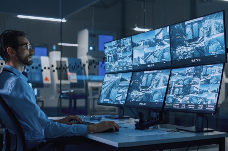Industry 4.0 Modern Factory: Security Operator Controls Proper Functioning of Workshop Production Line, Uses Computer with Screens Showing Surveillance Camera Feed. High-Tech Security